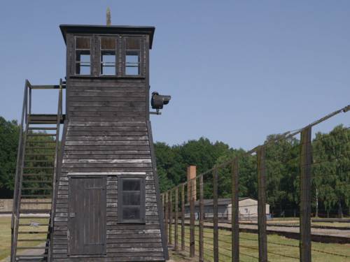 Zu sehen ist ein Wachturm aus dunklem Holz, der neben einem Stacheldrahtzaun steht.